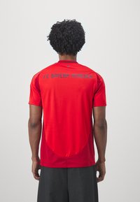 Rød atletisk trøye med korte ermer, med "FC BAYERN MÜNCHEN" i blå skrift på ryggen, designet med en glatt tekstur og runding i bunnen.
