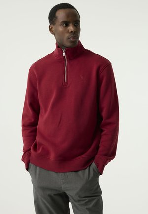 Man wearing a red quarter-zip sweatshirt with hands in gray pants pockets, standing and looking to the side against a plain background.
