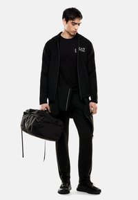 Completo sportivo nero composto da un hoodie con zip, una maglietta e pantaloni aderenti con dettagli riflettenti. Lo zaino è anch'esso nero con chiusure a zip.
