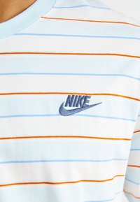 Medvilninis marškinėliai su šviesiai mėlynomis, baltomis ir oranžinėmis horizontaliomis juostelėmis. Papuoštas mėlynu Nike logotipu kairėje krūtinės pusėje.
