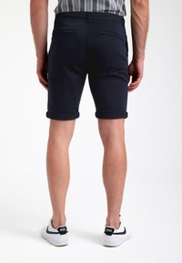 Pantalones cortos azul marino con puños enrollados, hechos de una tela ligera. Presentan dos bolsillos traseros y una textura suave; se llevan con zapatillas blancas.