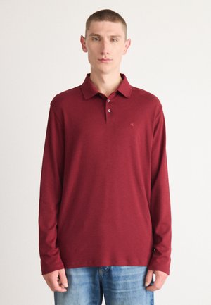 Burgunderfarvet polo med lange ærmer, tre-knaps placket, blød tekstur, diskret logo på brystet og klassisk kravedesign. Båret sammen med blå jeans.