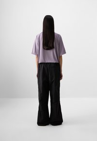 Une personne se tient de dos face à la caméra, portant une chemise lavande et un pantalon noir à jambes larges, sur un fond gris minimaliste.