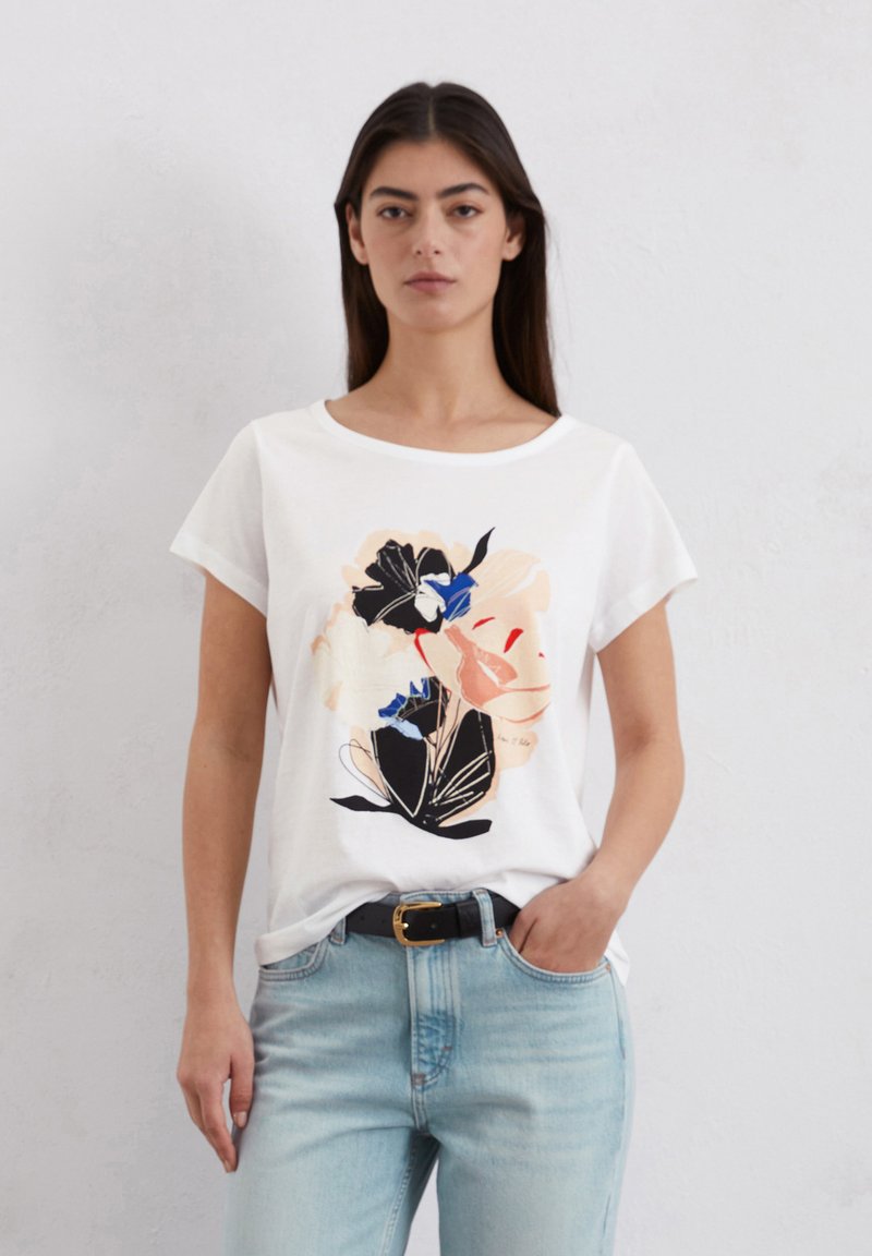Witte katoen t-shirt met een bloemendessin in perzik, zwart en blauw. Korte mouwen en een ronde halslijn, gecombineerd met lichtblauwe jeans.