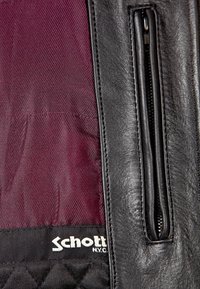 Gros plan sur un doublure en tissu bordeaux avec l'étiquette Schott N.Y.C. à côté d'une poche zippée en cuir noir.