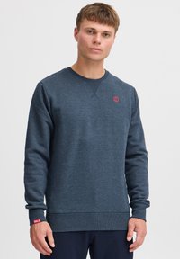 Sudadera azul marino de tejido suave con cuello redondo, puños y dobladillo acanalados, que presenta un pequeño logo rojo en el pecho y una etiqueta en la manga.