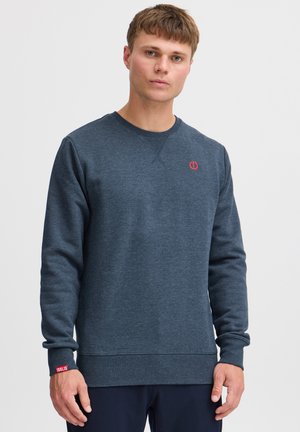 Sudadera azul marino de tejido suave con cuello redondo, puños y dobladillo acanalados, que presenta un pequeño logo rojo en el pecho y una etiqueta en la manga.