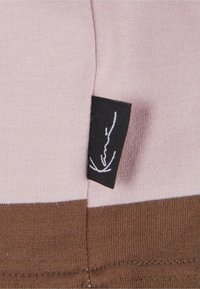 Bland av bomullsblanding med en rosa nyanse og brune horisontale striper. Har en svart etikett med hvit signaturdetalj.