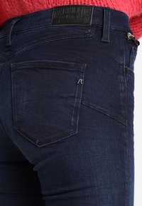 Vaqueros de denim azul oscuro con un diseño ajustado, costuras visibles y una etiqueta en la cintura. Un solo bolsillo trasero con un acento metálico.