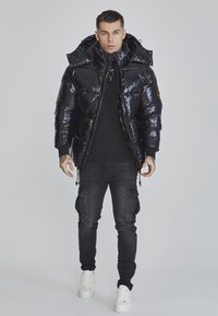 Veste en duvet noire avec une finition brillante, fermeture éclair à l'avant et capuche à cordon. Portée avec un pantalon cargo noir et des baskets blanches.