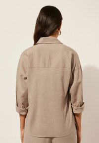 Camisa de lino beige con un corte relajado, mangas enrolladas y cuello caído; textura sutil y dobladillo trasero recto.
