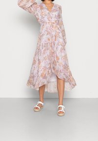 Blommig midiklänning i ljus lavendel med långa ärmar, omslagdesign och ankellång fåll med orange och rosa blommönster. Vita sandaler.