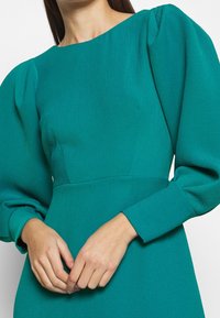 Robe bleu sarcelle avec manches longues bouffantes, tissu texturé et encolure bateau. Comprend un corsage cintré et des détails de jupe lisse.