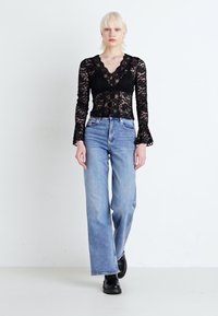 Μαύρο δαντελωτό κορμάκι με μακριά μανίκια και V-λαιμόκοψη, σε συνδυασμό με ψηλόμεσα ανοιχτό μπλε τζιν και μαύρες μπότες μέχρι τον αστράγαλο, μπροστά σε λευκό φόντο.