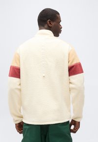 Άνδρας με γυρισμένη την πλάτη φοράει κρεμ φλις μπουφάν με μπεζ και κόκκινες ρίγες στα μανίκια και σκουροπράσινο παντελόνι, σε απλό φόντο.