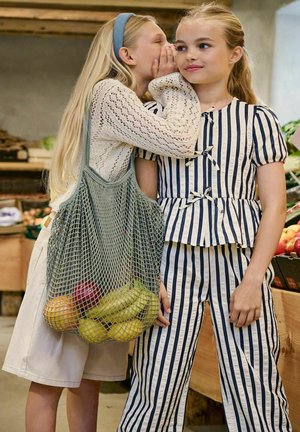 Twee meisjes op een markt, de een fluistert tegen de ander, die een gestreepte outfit draagt; het fluisterende meisje draagt een netzak met fruit.