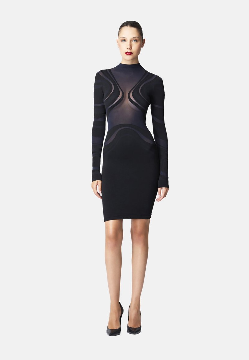 Donna che indossa un vestito nero aderente al ginocchio con motivi geometrici trasparenti e opachi, abbinato a tacchi neri alti, in piedi su uno sfondo semplice.