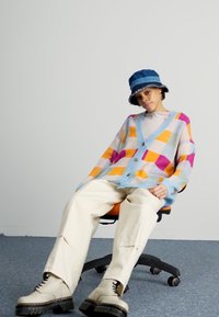 Gros cardigan coloré avec des accents bleus, présentant un motif géométrique en orange, violet et teal. Associé à un pantalon crème et des bottes chunky.