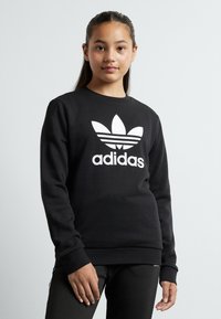Juodas džemperis ilgomis rankovėmis, su dideliu baltu Adidas logotipu ir trimis juostelėmis ant priekio, megztomis rankogaliais ir apatine dalimi.