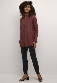 Camicia di raso color marrone bordeaux con abbottonatura frontale e maniche lunghe, abbinata a jeans skinny neri strappati e sandali neri con tacco a blocco.