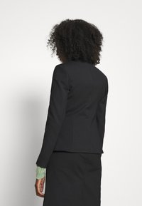 Persona con capelli ricci che indossa un blazer nero aderente sopra una maglietta verde a maniche lunghe, vista di spalle su uno sfondo semplice.