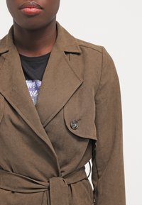 Brą Coat w kolorze brązowym, z teksturą, o dopasowanym kroju, dużym kołnierzu i zapięciu na guziki. Posiada pasek w talii oraz delikatny połysk.