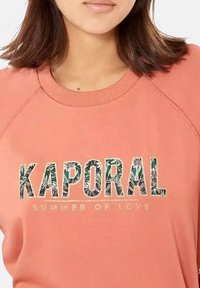 Sweatshirt rose avec "KAPORAL" en motif floral texturé et "SUMMER OF LOVE" en doré, avec un col rond et des manches raglan.