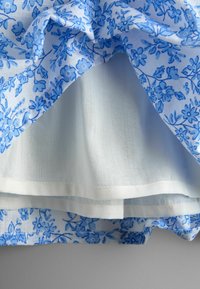 Blau gemustertes Stoff mit Blumenmuster, zurückgefaltet, um den darunter liegenden einfarbigen weißen Stoff zu zeigen, der geschichtete Textilstrukturen und Nähte offenbart.