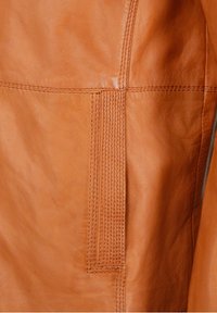 Læderjakke i tan nærbillede med glat tekstur, kontrasterende syninger og en dekorativ søm med en lodret ribbet detalje.