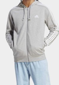 Felpa con cappuccio grigia con cerniera, realizzata in tessuto morbido, con dettagli a tre strisce bianche sulle maniche e logo adidas sul petto.