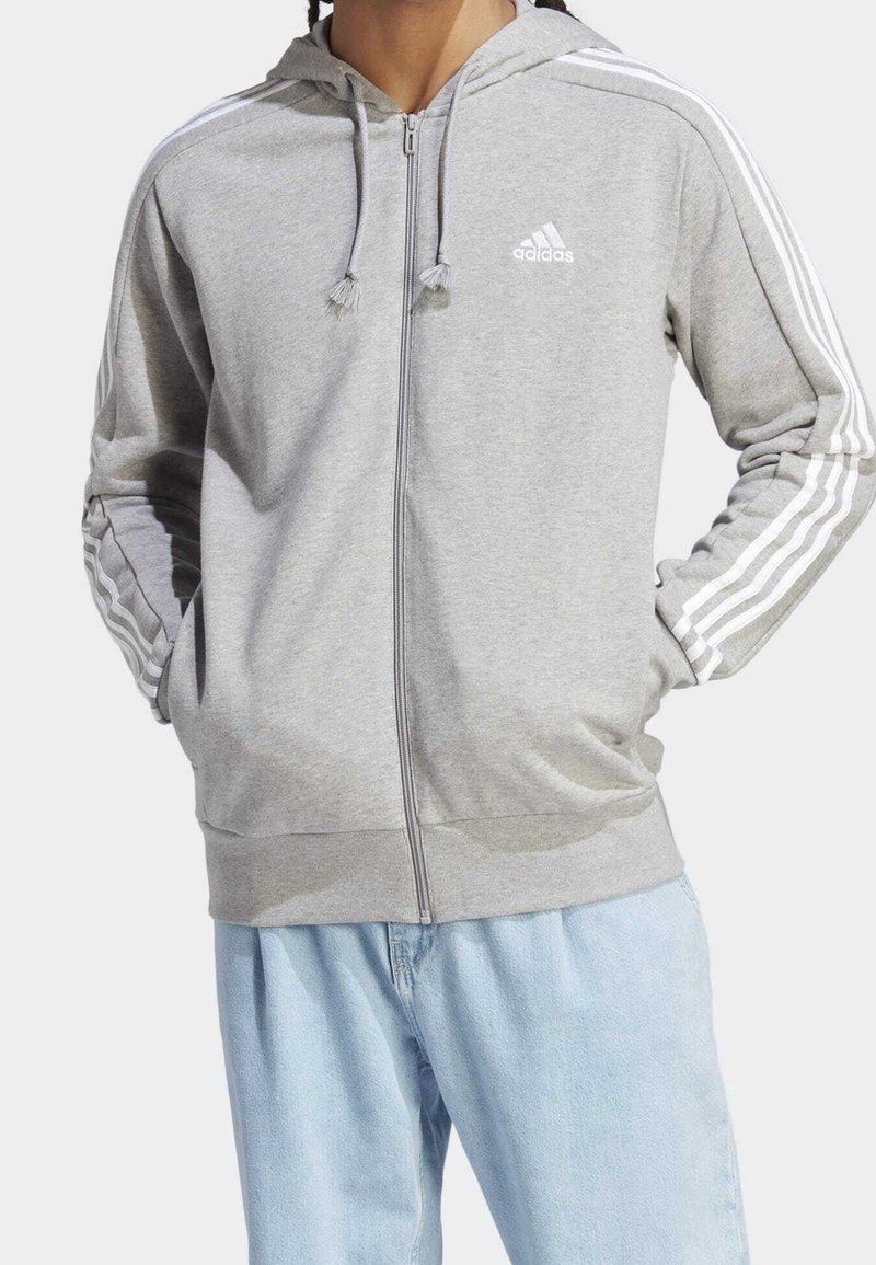 Felpa con cappuccio grigia con cerniera, realizzata in tessuto morbido, con dettagli a tre strisce bianche sulle maniche e logo adidas sul petto.