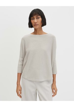 Vrouw met kort zwart haar, gekleed in een loszittende lichtbeige top met lange mouwen en lichtgekleurde broek, staande tegen een effen achtergrond.