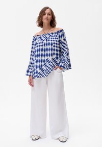 Bluzka bez ramiączek w niebiesko-białym wzorze ikat, z luźnymi rękawami i swobodnym krojem, w połączeniu z białymi spodniami o szerokich nogawkach i płaskimi butami.