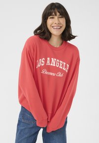 Sweatshirt rouge avec le texte blanc "LOS ANGELES Dreamers Club", présentant une coupe ample et des poignets côtelés, associé à un jean bleu.