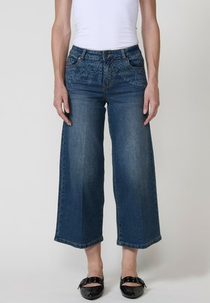Vaqueros anchos de cintura alta en denim azul oscuro con bordado floral en la cinturilla. Acabados con un dobladillo sin terminar. Zapatos planos negros.
