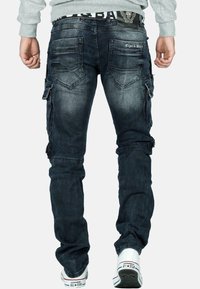Jeans cargo bleu foncé avec plusieurs poches, coupe fuselée et légères décolorations. Présente une ceinture de marque et des détails de couture visibles.