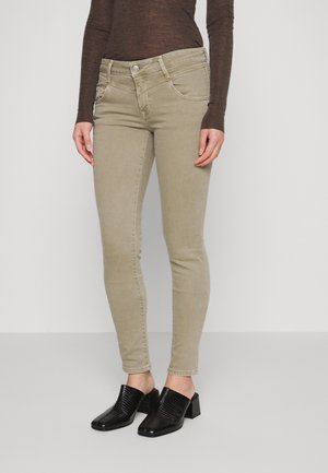Femme portant un pantalon beige ajusté, un haut à manches longues marron foncé et des chaussures à talons noires sans lacets, debout devant un fond blanc.