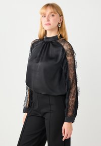 Schwarze Bluse mit langen Spitzenärmeln, hohem Kragen und glattem Satin-Korpus. Spitzenakzente verleihen der Gestaltung Textur und Detail.