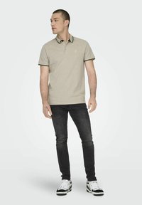 Lichtbeige polo shirt met zwarte accenten en een klein logo, gecombineerd met donkere jeans en zwart-witte sneakers. Man staat zijwaarts.