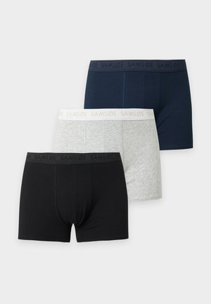 Jeune homme en forme debout pieds nus en boxer noir sur un fond clair uni, regardant vers sa gauche.