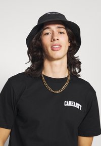 Chapeau seau noir avec logo blanc, t-shirt noir avec nom de marque blanc, et un épais collier en chaîne dorée. Fond uni.