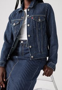 Tummansininen farkkutakki, jossa valkoiset pystysuorat raidat, napit edessä, kaksi etutaskua ja klassinen kaulus, yhdistetty samaa kangasta olevaan hameeseen.