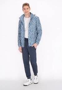 Blauwe lichtgewicht jas met een vispatroon, gecombineerd met navy joggers. Een wit T-shirt eronder en witte sneakers maken de outfit compleet.