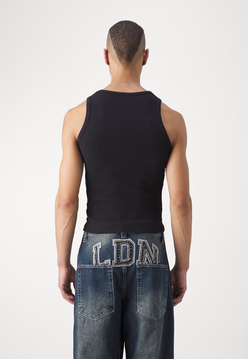 Muž s unikátním oholeným účesem nosí černý tílko bez rukávů a volné modré džíny s velkými písmeny "LDN" na zadních kapsách.