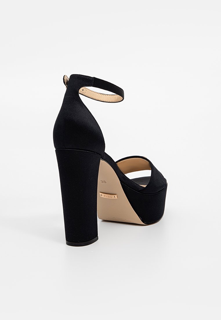 Talons compensés en suède noir avec un large talon haut, une sangle à la cheville et un bout rond. La semelle est beige et présente une légère courbure. Taille 38.