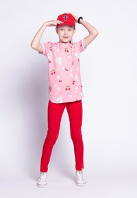 Różowa koszulka z krótkim rękawem w wzory wiśni i kwiatów, zestawiona z obcisłymi czerwonymi legginsami i białymi sneakersami. Czerwona czapka z logo "T".