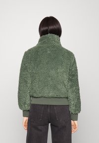 Texturerad grön fleecejacka med hög krage, ribbade ärmslut och nederkant, samt en avslappnad passform. Materialet verkar mjukt och varmt, lämpligt för kallt väder.
