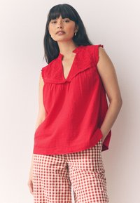 Rote Bluse mit besticktem Kragen und gerafften Rändern, kombiniert mit rot-weiß karierten Gingham-Hosen. Weicher, strukturierter Stoff.
