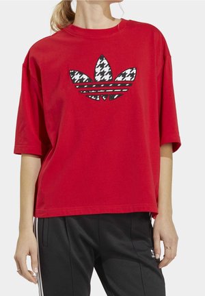 Mujer vistiendo una camiseta roja holgada con un logo en blanco y negro y pantalones de chándal negros de Adidas con rayas blancas.