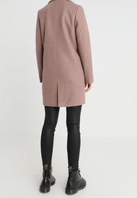 Lang skræddersyet frakke i pastelfarvet uldblanding, med lige snit, stort krave og centralt rygsyg. Båret over sorte tætsiddende bukser og støvler.